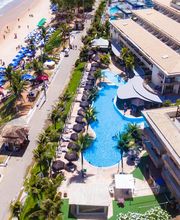 Vista aérea de um resort à beira-mar com piscina, palmeiras e guarda-sóis na praia.