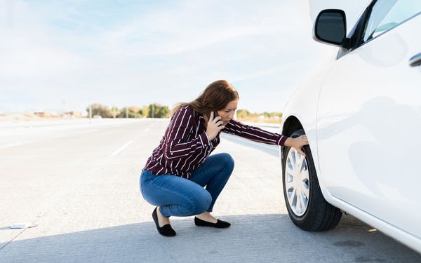 Frau kniet neben Reifen ihres Autos und ruft den Abschleppdienst, da sie nicht mehr weiterfahren kann.
