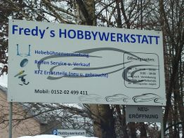 Schild: Fredy's Hobbywerkstatt mit Öffnungszeiten und Kontaktnummer unter Bäumen.