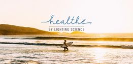Surfer holding a board, wading in sunlit waves at sunset; text: "healthe by Lighting Science".