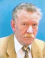 Älterer Mann mit Schnurrbart in einem beigen Anzug vor blauem Hintergrund.