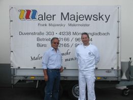 Zwei Männer vor einem Anhänger mit der Aufschrift "Maler Majewsky" und Kontaktdetails.