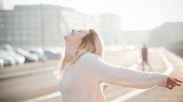 Woman in white sweater enjoys sunlight with outstretched arms on a street.
