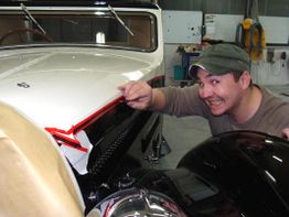 Mann lächelt neben einem Oldtimer in einer Werkstatt. Er zeigt auf das Auto mit rotem Klebeband.