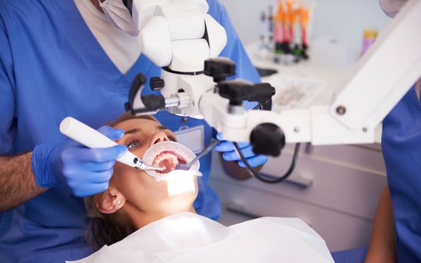 Eine junge Frau liegt mit geöffnetem Mund auf einem Behandlungsstuhl. Ein Zahnarzt behandelt ihre Zähne.