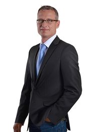 Ein Mann im Anzug mit blauer Krawatte steht auf weißem Hintergrund.