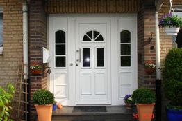 Weiße Haustür mit Fenstern und umgeben von Blumenkübeln und Ziegelwänden.