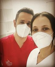 Dos personas con mascarillas, una con uniforme rojo y otra con blanco, en un entorno clínico.