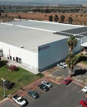 Vista aérea de una gran nave industrial blanca con estacionamiento y palmeras alrededor.