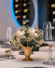 Centro de mesa floral en un jarrón dorado, rodeado de copas de vino en un restaurante elegante.