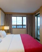 Quarto de hotel com cama de casal, roupa de cama vermelha, janela grande e TV na parede.