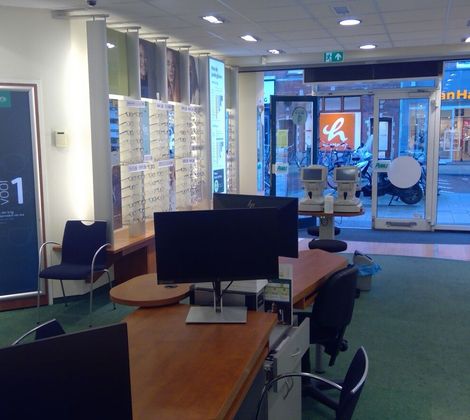 Optiekwinkelinterieur met stoelen, display met brillen en een bureau met computers.