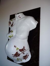 Weiße Gipsfigur eines schwangeren Torsos mit braunen und grünen Blumenmustern an der Wand.