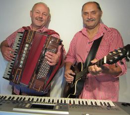 Zwei Männer in karierten Hemden spielen Akkordeon und Gitarre, ein Keyboard steht vor ihnen.