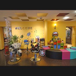 Ein bunter Friseursalon mit Spielzeugmotorrädern und farbenfrohen Sitzecken vor gelber Wand.