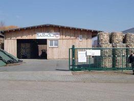 Holzlagerhalle mit gestapelten Holzpaletten, offenes Tor und Firma "Saxholz" am blauen Himmel.