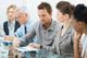 Five people in a business meeting, discussing and taking notes around a glass table.