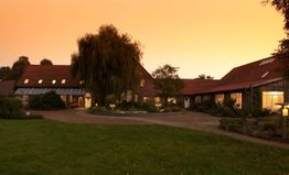 Großer Bauernhof bei Sonnenuntergang mit beleuchteten Fenstern und einem Baum im Vordergrund.