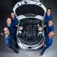 Six mechanics smiling around a car with its hood open, showcasing the engine.