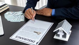 Man signing insurance document, with cash and house model on the desk.