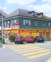 Straßenansicht eines Gebäudes mit Pizzeria und Kebap-Laden, mehrere Autos parken davor.