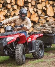 Persona en un ATV rojo traslada leña en un remolque frente a una gran pila de troncos.