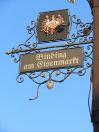Metallisches Schild mit der Aufschrift "Binding am Eisenmarkt" und Adleremblem vor blauem Himmel.
