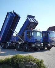 Zwei blaue Lastwagen mit gekippten Ladeflächen auf einem Parkplatz bei sonnigem Wetter.