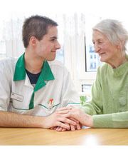 Ein Pfleger hält die Hand einer älteren Frau, beide lächelnd an einem Tisch sitzend.