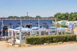 Blaues Café mit Sonnenschirmen draußen, umgeben von Wohnwagen und grünen Bäumen.