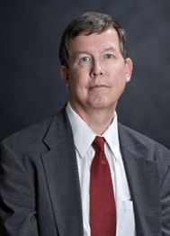 Man in a suit and red tie with a neutral expression, dark background.