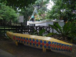 Escultura colorida de cobra em parque com vegetação e barco ao fundo.