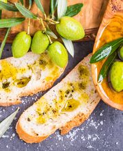 Pane con olio d'oliva, olive verdi e un cucchiaio di legno su un tavolo grigio.