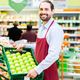 Smiling grocery worker in a red apron holding a box of green apples in a supermarket.