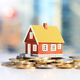 A model house sits on a stack of coins against a blurred city background.