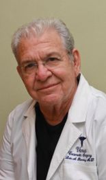 Elderly man in a white lab coat with embroidered text, smiling gently against a neutral background.