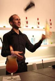 Barkeeper jongliert mit Shaker-Flasche, brennender Cocktail im Vordergrund, Bar im Hintergrund.