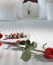 Cama con pétalos de rosa, fresas cubiertas de chocolate y una rosa roja sobre el edredón blanco.