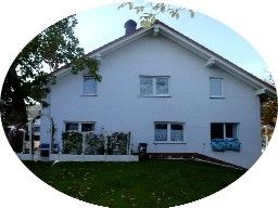Weißes Haus mit Garten, Bäumen und einem kleinen Zaun im Vordergrund.