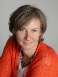 Eine Frau mit rotem Pullover lächelt in die Kamera vor grauem Hintergrund.