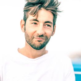 Un homme avec des cheveux ébouriffés et barbe, portant un t-shirt blanc, sourit légèrement.