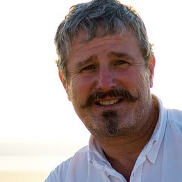 Ein lächelnder Mann mit Schnurrbart und Bart, trägt ein weißes Hemd, bei hellem Hintergrund.