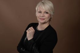 Frau mit kurzem, blondem Haar und schwarzer Jacke, posiert vor braunem Hintergrund.