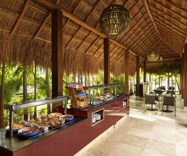 Buffet tropical bajo techo de palapa, mesas al fondo rodeadas de vegetación exuberante.