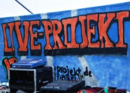 Graffiti-Wand mit Schriftzug "LIVE-PROJEKT" in Orange und Blue, davor Audio-Equipment.