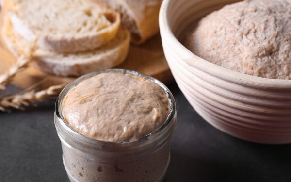 Ein Sauerteigstarter in einem Glas. Im Hintergrund befindet sich ein Sauerteig in einem Gärkörbchen, daneben liegt ein fertig gebackenes Sauerteigbrot.