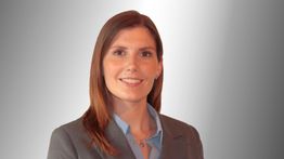 Eine Frau in Business-Outfit vor einem grauen Hintergrund, lächelnd in die Kamera.