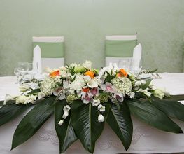 Centrotavola floreale con foglie verdi e fiori bianchi e arancioni su una tavola bianca.