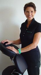 Frau in schwarzem Polohemd lächelt, steht neben einem schwarzen Massagestuhl.