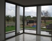 Vista externa através de janelas de vidro em sala, com árvores e construção ao fundo.